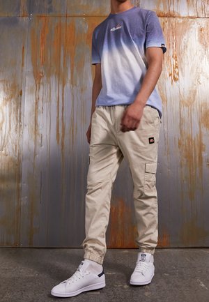 Camiseta de color azul claro con degradado y mangas cortas, combinada con pantalones cargo beiges con puños elásticos y bolsillos de parche, llevados con zapatillas blancas.