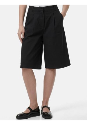 Person iført sorte, knælange, skræddersyede shorts med sidelommer og sorte Mary Jane-stil sko på en hvid baggrund.