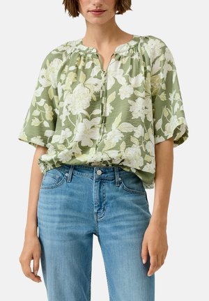 Vrouw draagt een lichtgroene blouse met bloemenprint en halflange mouwen, ingestopt in medium-was blauwe spijkerbroek met knopen aan de voorkant en zakken.