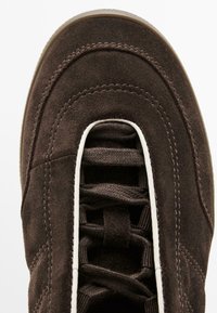 Primo piano della punta di una scarpa in suede marrone scuro con lacci abbinati, bordo bianco attorno all'area dei lacci e cuciture dettagliate nella parte superiore.