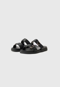 Svarta sandaler med två breda remmar, prydda med en guldfärgad cirkelförsköning. Strukturerad sula och platt design för komfort.