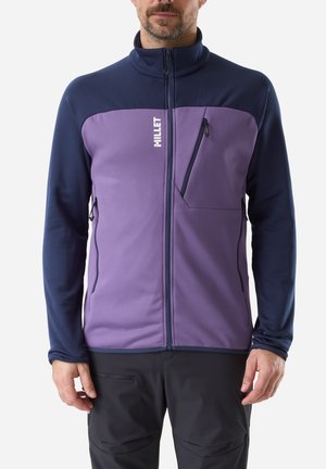 Man wearing a purple and navy zip-up jacket with a high collar and zippered chest and side pockets, standing against a white background.