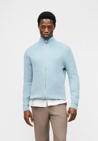 Άνδρας που φοράει ανοιχτό μπλε πουλόβερ με φερμουάρ, άσπρο πουκάμισο με ρίγες και μπεζ παντελόνι, στέκεται μπρ οστά σε απλό λευκό φόντο.