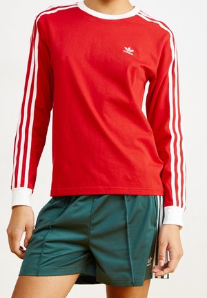 Personne portant un t-shirt rouge à manches longues Adidas avec des rayures blanches et un short vert Adidas avec des rayures blanches sur les côtés, debout sur un fond uni.
