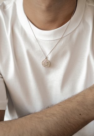 Person wearing a silver round pendant necklace with a rose design, dressed in a white crew-neck t-shirt, arm partially visible.