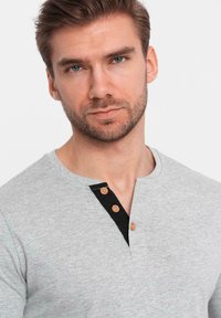 Grå långärmad skjorta med en svart halsringning och tre bruna knappar, mjuk textur och rundad hals. Ingen mönstring eller accenter.