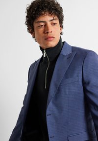 Blue checkered blazer with structured lapels, worn over a fitted black turtleneck, featuring a zipper and ribbed texture.