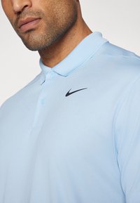 Polo de cor azul clara feito de tecido suave, com colarinho abotoado, apresentando o logótipo preto da Nike na área do peito esquerdo.