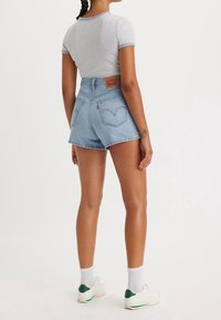 Shorts di jeans blu a vita alta con un'etichetta in pelle del brand, abbinati a una maglietta grigia e sneaker bianche con accenti verdi.