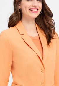 Oranges Blazer mit einer strukturierten Oberfläche, mit Reverskragen und zwei Knöpfen.getragen über einem passenden orangen Oberteil mit einem subtilen Glitzereffekt.