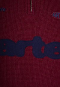 Maglione rosso lavorato a maglia con colletto a metà zip, caratterizzato da un ampio design grafico blu navy che incorpora lettere e forme.