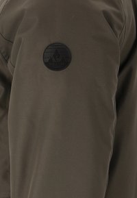 Oliven grøn jakke med en glat, mat overflade. Har et sort cirkulært logo mærke på ærmet. Funktionelt design med minimal detaljer.