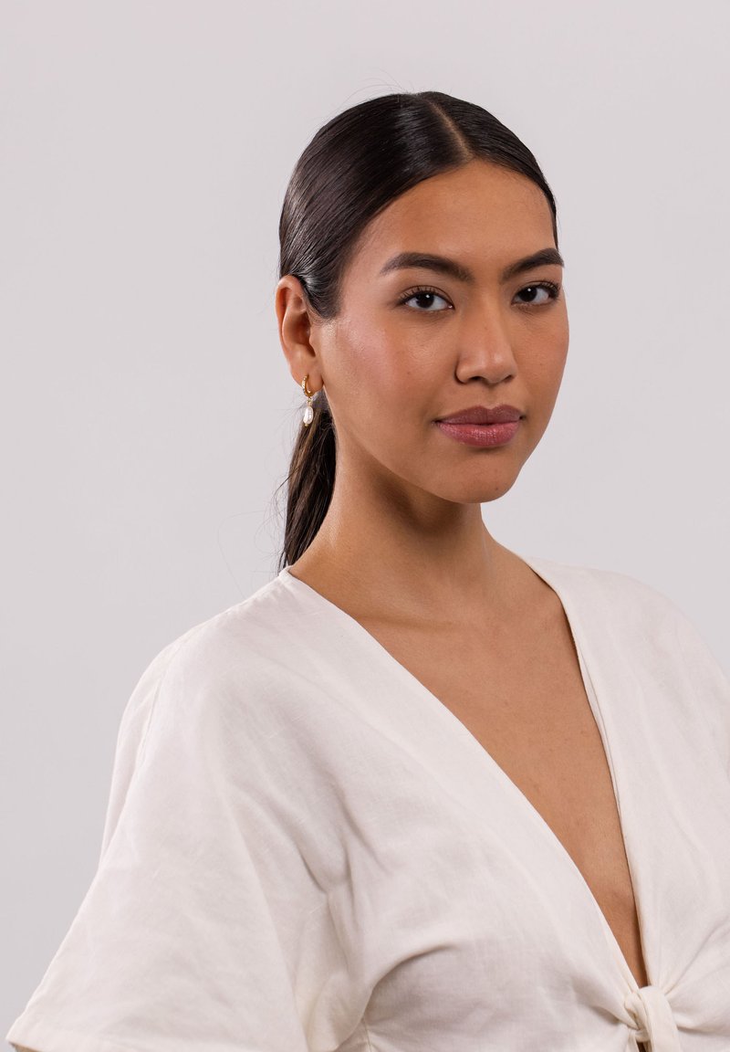 Eine Frau trägt eine weiße, leinen Wrap-Bluse mit tiefem V-Ausschnitt und einer Knoten-Details in der Taille. Sie hat goldene Creolen mit einer Perlenakzent.