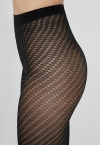 Černé punčochy s vzorem vln a oválnými výřezy. Hladký materiál s širokým černým pasem pro podporu.