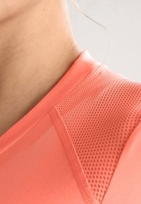 Korallenfarbenes Sportshirt aus glattem Stoff mit strukturierten Netzpaneelen an der Schulter, die Atmungsaktivität und Komfort betonen.