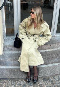 Beige trench coat with a belted waist, large collar, and button details. Worn with black pointed shoes and sheer black tights. Black handbag beside.