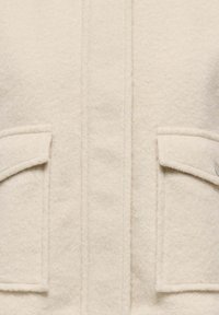 Beige uldblandet jakke med en blød tekstur, to forlommer med klapper og knapper langs knaplisten.