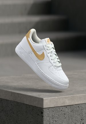 Bílé kožené tenisky se zlatými akcenty, s perforovanou špičkou, polstrovaným límcem a strukturovanou podešví, suspendované nad betonovým povrchem.