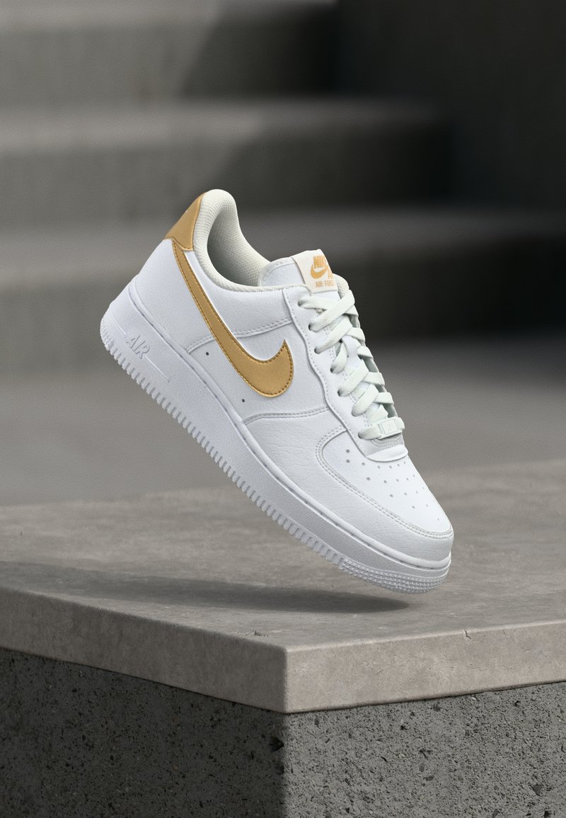 Bijele kožne tenisice s zlatnim akcentima, s perforiranim prstom, podstavljenim okovratnikom i teksturiranim potplatom, suspendirane iznad betonske površine.