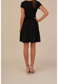 Robe courte noire à manches courtes avec une ceinture nouée à la taille et une jupe évasée. Le tissu texturé présente un motif subtil. Le modèle porte des sandales à talons hauts à lanières.