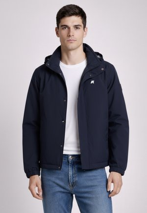 Giovane uomo con una giacca a cappuccio blu navy sopra una t-shirt bianca e jeans blu, in piedi contro uno sfondo chiaro e semplice.