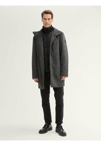 Abrigo de mezcla de lana gris con una capa interior con cremallera, capucha ajustable y bolsillos laterales, llevado sobre pantalones oscuros y zapatos negros de cordones.