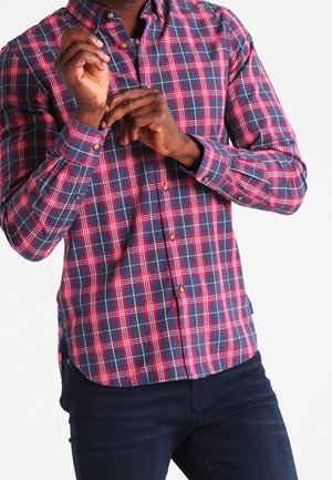 Mężczyzna zapinający mankiet koszuli w czerwoną, niebieską i białą kratę, ubrany w ciemnoniebieskie dżinsy, na białym tle.