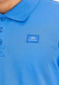 Zils polo krekls, kas izgatavots no teksturēta auduma, ar apkakles dizainu, trim pogām un "Alpha Industries" logo uz krūtīm.