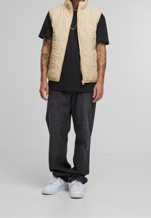 Ljusbeige kviltad väst över en svart t-shirt, kombinerad med mörkgrå, löst sittande byxor och vita sneakers. Minimalistisk design med dragkedjedetaljer.