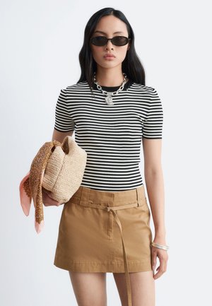 Femme portant des lunettes de soleil noires, un t-shirt à manches courtes rayé noir et blanc, une jupe beige, un collier et un bracelet en argent, tenant un sac en paille avec un foulard.
