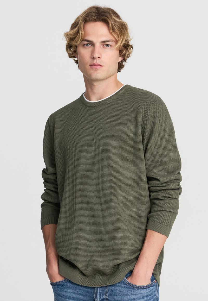 Olive zielony, teksturowany długi rękaw sweter z okrągłym dekoltem. Posiada luźny krój i ściągacze przy mankietach. Połączony z niebieskimi dżinsami.