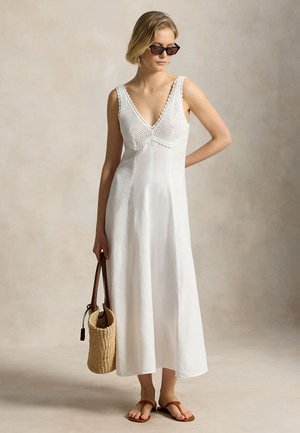 Kvinde i ærmeløs hvid kjole med strikket krop, iført solbriller, holder en vævet taske og har brune sandaler på.