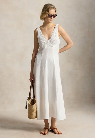 Woman in sleeveless white dress with knitted bodice, wearing sunglasses, holding a woven bag and wearing brown sandals.