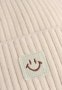 Beanie a coste rosa chiaro con una toppa bianca testurizzata che presenta un sorriso ricamato marrone.