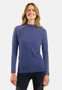 Langärmliges Performance-Oberteil aus strukturiertem, dunkelblauem Stoff, mit hohem Kragen und tailliertem Design, kombiniert mit konturierten schwarzen Leggings.