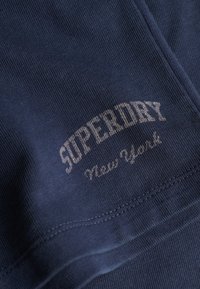 Ciemnoniebieska tkanina z białym przeszywanym napisem "SUPERDRY New York" oraz widocznymi szwami.