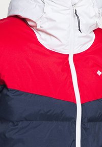 Padded Jacke mit einer weißen Kapuze; verfügt über einen roten oberen Bereich, einen marineblauen unteren Bereich und einen weißen Reißverschluss. Strukturierter Stoff mit kontrastierenden Farben.