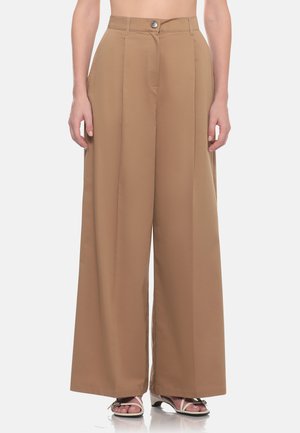 Pantaloni beige a vita alta e gambe larghe con pieghe frontali indossati da una persona in piedi con sandali bianchi open toe.