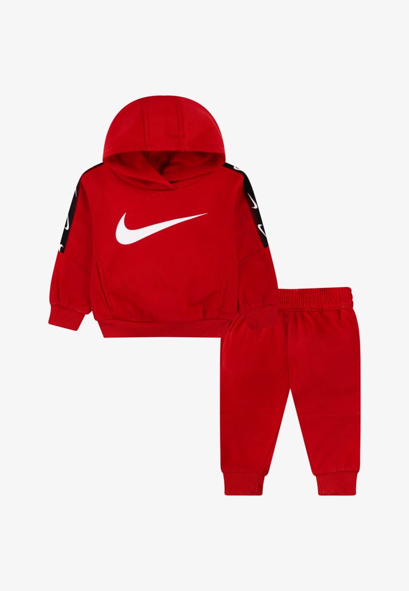 Punainen taaperon urheiluasu, jossa on huppari ja elastiset housut, joissa on valkoiset Nike-logot rinnassa ja hihoissa.