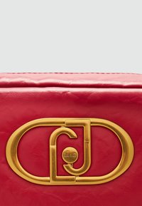 Rote Ledertasche mit einem auffälligen goldfarbenen Logo-Verschluss, glatter Oberfläche und strukturiertem Design mit sichtbaren Nähdetails.
