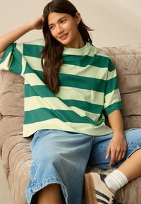 Tricou supradimensionat cu dungi verzi și verde deschis, realizat dintr-un material moale, asortat cu pantaloni scurți din denim albastru deschis. Șezând pe o canapea bej.