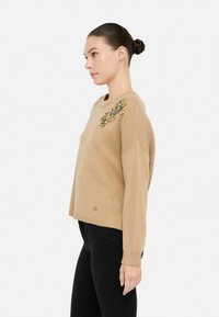 Maglione lavorato a maglia beige con scollatura a girocollo, caratterizzato da un'abbellimento decorativo di pietre gialle e grigie sulla spalla.