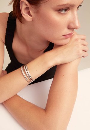Trois jolis bracelets en argent lisse empilés sur un poignet, sur fond neutre. Le bras présente un teint de peau naturel et légèrement texturé.