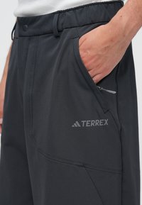 Tumši pelēkas Terrex bikses ar gludu faktūru, elastīgu jostasvietu, rāvējslēdzēja kabatu un atstarojošu logo uz augšstilba.