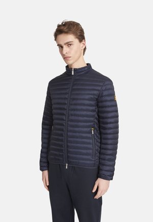 Junger Mann trägt eine marineblaue gesteppten Jacke zum Reißverschluss mit Reißverschlusstaschen, steht vor einem einfarbigen weißen Hintergrund.