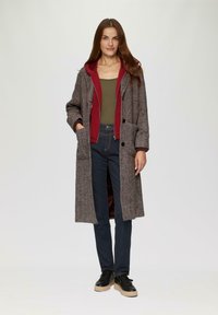Lang jasje met een visgraatpatroon in bruin en grijs, met een bordeauxrode capuchon. Gepaarde met een getailleerd groen shirt en donkere jeans. Zwarte sneakers.