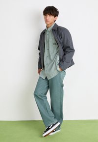 Model wearing a gray jacket over a green and white striped shirt, paired with light green pants and black sneakers with white accents.