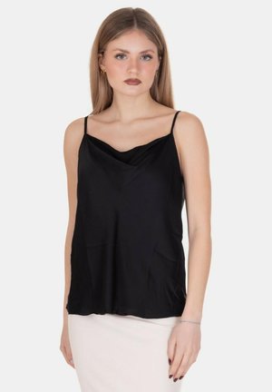 Giovane donna con lunghi capelli biondi che indossa un top nero senza maniche con sottili spalline e una gonna bianca, in piedi davanti a uno sfondo semplice.