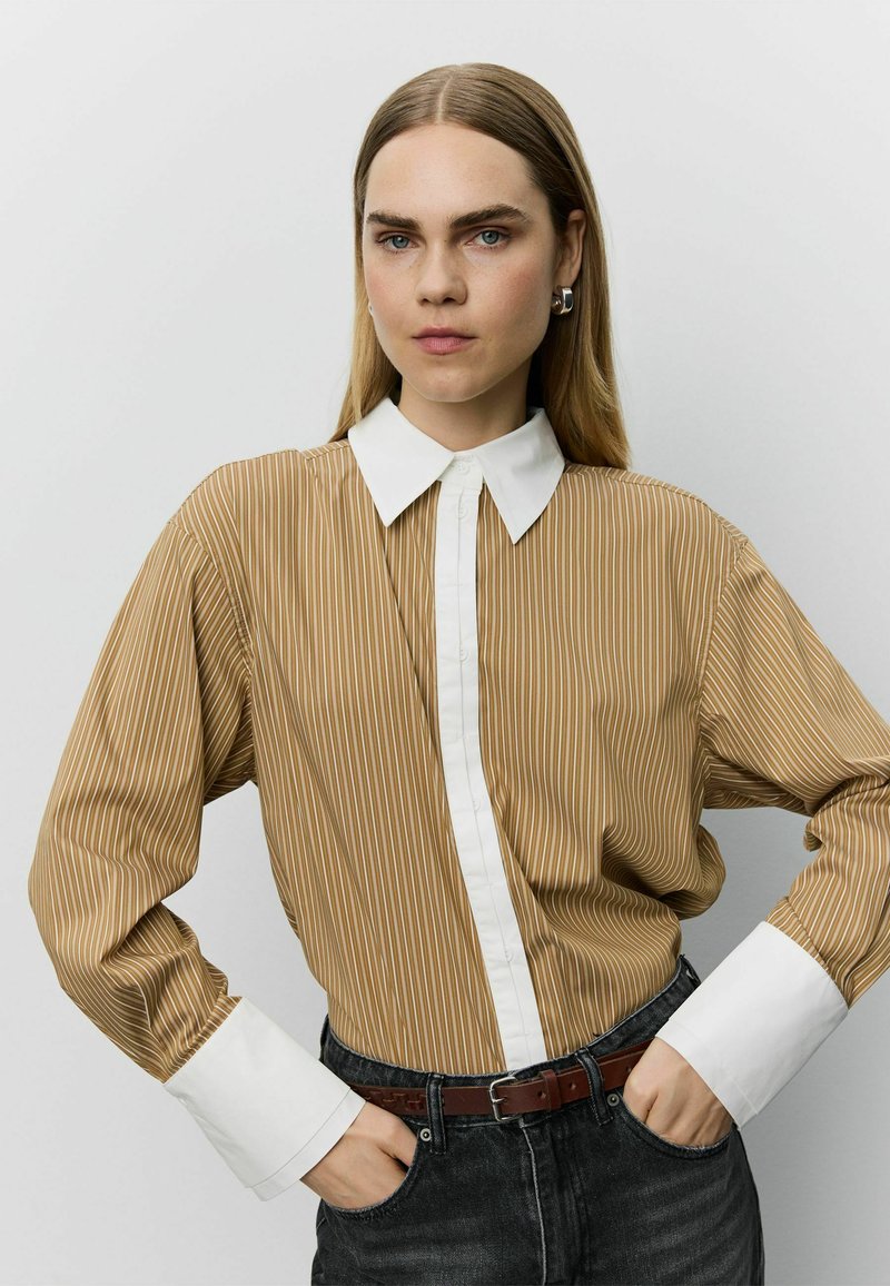 Femme aux cheveux blonds raides portant une chemise marron rayée avec col et poignets blancs, jean noir et boucles d'oreilles argentées en anneau, mains dans les poches.