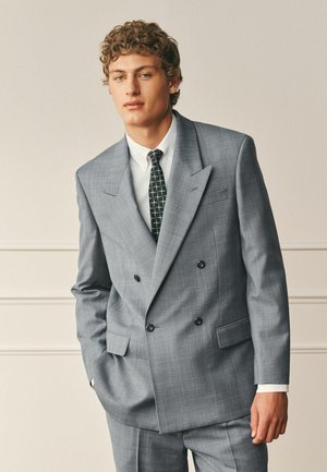 Junger Mann mit lockigem Haar, der einen hellgrauen Doppelreiher-Anzug, ein weißes Hemd und eine schwarze gemusterte Krawatte trägt, steht mit einer Hand in der Tasche.